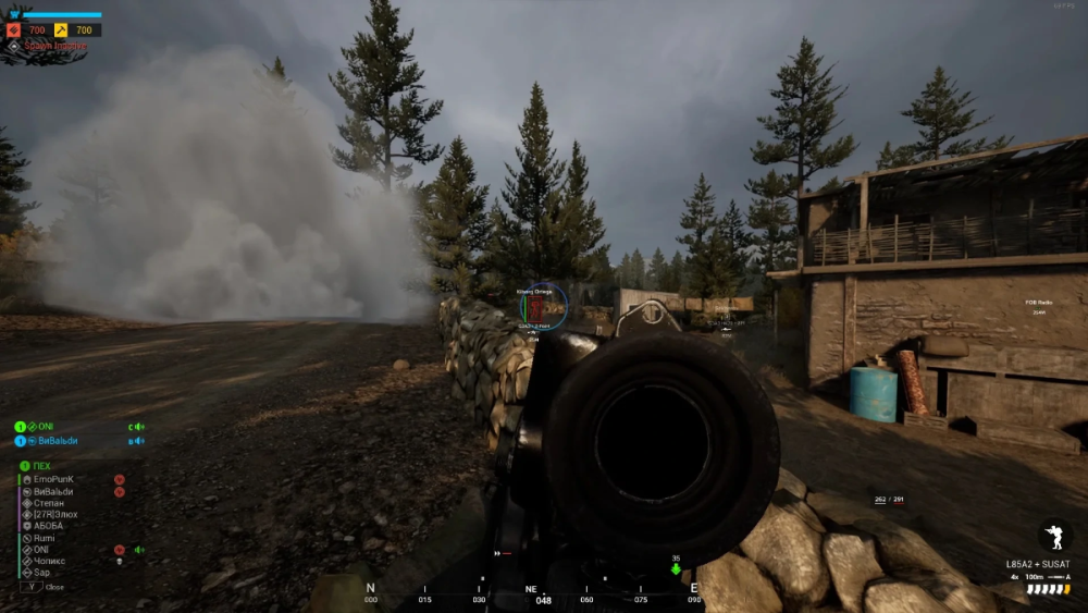 First-person shooter game scene showing a dark rifle aimed at a dirt road, flanked by trees and a stone wall. Smoke billows on the left, adding tension.