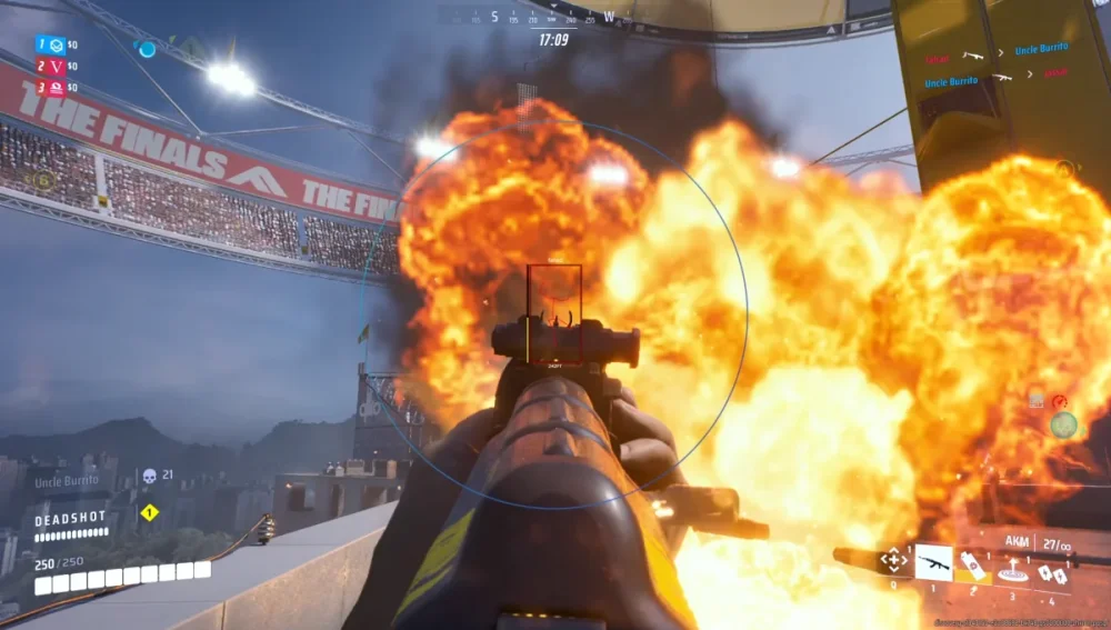 First-person shooter game scene showing a weapon sight aimed at a large explosion. Bright flames dominate with a stadium in the background, intense action.